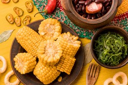 Pratos típicos de festa junina com milho cozido, feijão com linguiça e acompanhamentos coloridos sobre mesa decorada.