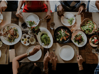 A imagem mostra uma mesa de restaurante vista de cima, com várias pessoas reunidas compartilhando uma refeição. A mesa está cheia de pratos variados, incluindo massas, saladas, carnes, vegetais e acompanhamentos, sugerindo uma refeição sofisticada e diversa.