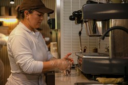 Una persona utiliza una máquina de café espresso en una cocina industrial, mientras lleva guantes de plástico, una camisa blanca y un delantal a rayas. En el fondo, se observan utensilios de cocina y tazas apiladas.