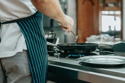 Um cozinheiro usando um avental listrado, segurando uma espátula enquanto frita algo em uma panela, em uma cozinha moderna