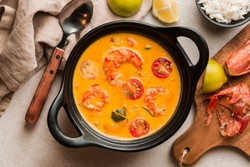 Un recipiente negro con sopa de camarones, en un caldo de color amarillo anaranjado. La sopa contiene camarones, rodajas de tomate y otros ingredientes. 