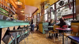 Imagem de salão de restaurante, estilo gastrobar, com um homem sentado na banqueta do balcão enquanto a mesa ao seu lado é ocupada com um grupo de quatro pessoas.