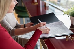 Braço de uma mulher assinando um documento com outra mulher ao seu lado.