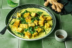 Prato de tofu grelhado com espinafre em um molho cremoso amarelo, servido em um prato com fundo verde, acompanhado por gengibre fresco