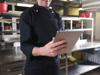 En la imagen, un chef viste un uniforme negro y sostiene una tableta con ambas manos. Parece estar consultando una receta o revisando información digital.