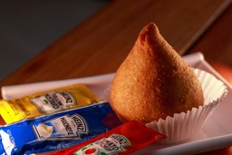 Coxinha em um prato com sachês de ketchup, maionese e mostarda.