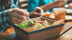 Close em primeira pessoa de uma pessoa utilizando um garfo para comer uma salada colorida servida em uma caixa de papelão kraft.