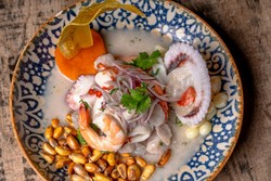 Un plato típico de Semana Santa en Perú que muestra ceviche de mariscos servido en un plato decorativo con diseño azul y blanco. Los ingredientes incluyen camarones, vieiras, cebolla roja en rodajas, cilantro, maíz tostado y choclo. El plato está acompañado por una rodaja de camote y una pieza de plátano frito.