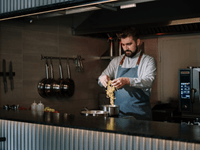 A imagem mostra um chef profissional preparando massa em uma cozinha moderna e bem equipada. Ele está concentrado em inserir a massa em uma panela sobre o fogão. O ambiente é limpo, organizado, e com utensílios pendurados na parede — típico de uma cozinha profissional eficiente, mas sem presença de clientes, o que remete diretamente ao conceito de dark kitchen.