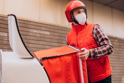 Logística de entrega: Motoboy vestido de colete laranja com uma blusa xadrez por baixo do colete, de capacete laranja e máscara com uma bag de entrega laranja em suas mãos saindo de um tubo branco que liga o restaurante a rua.