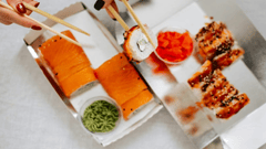 A imagem apresenta uma visão aérea em close-up de uma refeição de sushi para viagem, servida em embalagens de papelão prateadas sobre uma superfície clara.