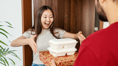 Uma jovem asiática aparece com uma expressão de extrema alegria e os braços abertos, comemorando a chegada da comida. Em primeiro plano, o entregador (visto de costas com uma camiseta vermelha) segura uma caixa de pizza que serve de apoio para duas embalagens brancas de viagem.