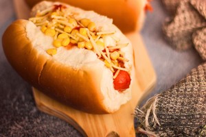 Completo brasileño con salchicha, maíz, papas fritas y salsa servido en pan suave sobre tabla de madera.