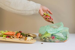 Uma mão colocando restos de vegetais em um saco plástico verde parcialmente cheio de resíduos orgânicos. Ao lado, há uma tábua de corte com mais restos de vegetais, como cenouras, cogumelos e folhas verdes.