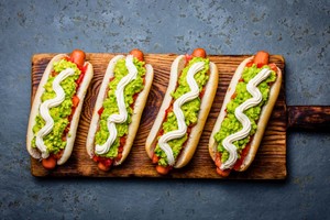 Cuatro completos con palta, tomate y mayonesa sobre tabla de madera, representando tipos de completos chilenos tradicionales.