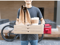 La imagen muestra a un repartidor sosteniendo varios artículos de comida para llevar. La persona, que viste una camiseta gris y jeans, lleva: Tres cajas de pizza apiladas. Dos bolsas de papel kraft, típicamente utilizadas para empaques de comida. Dos vasos de café o bebida caliente con tapas, dentro de un portavasos de cartón.