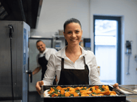 La imagen muestra a una mujer en una cocina profesional, en primer plano, sosteniendo una bandeja con alimentos preparados, probablemente verduras asadas, presentadas de manera ordenada y colorida. Ella viste una camisa blanca y un delantal oscuro, lleva el cabello recogido y sonríe directamente a la cámara, transmitiendo confianza, orgullo y profesionalismo.