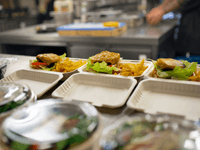 Imagem mostra marmitas sendo preparadas com hambúrguer, batatas fritas e saladas em uma cozinha profissional, limpa e organizada.