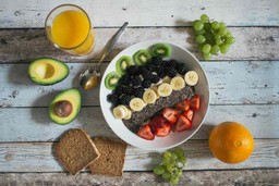 Bowl com frutas e chia, rodeado por uvas verdes, laranja, torradas, abacate e um copo de suco.