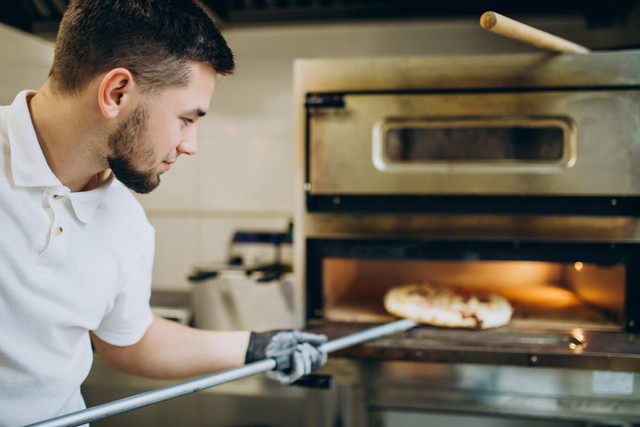 cafetería fantasma: Un joven sacando una pizza del horno, lleva una bata blanca y tiene el pelo corto.