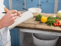 La imagen es un primer plano que se centra en las manos de una persona, probablemente un chef o cocinero, tomando notas en una cocina. La escena sugiere planificación o documentación de recetas.