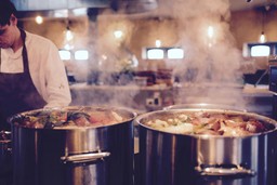 Grandes panelas no fogão com vapor, chef ao fundo, cena de cozinha de catering.