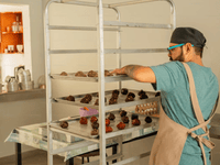 A imagem mostra um profissional em uma cozinha comercial organizando bandejas cheias de massas ou doces modelados (possivelmente brigadeiros ou cookies) em um carrinho de inox com várias prateleiras. Ele está usando avental, touca e óculos de proteção, demonstrando cuidado com a higiene e segurança no preparo dos alimentos.
