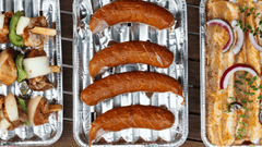 A imagem mostra uma vista aérea de três bandejas de alumínio descartáveis dispostas lado a lado sobre uma superfície de madeira escura, sugerindo preparo para churrasco ou grelhados.