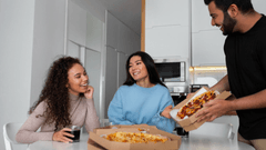 Duas mulheres estão sentadas à mesa branca sorrindo e conversando, enquanto um homem, em pé à direita, serve uma pizza saindo diretamente da caixa de entrega. Sobre a mesa, há outra caixa de pizza aberta e copos com um refrigerante escuro.