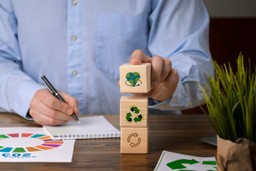 Cozinhas ecológicas: homem segurando bloquinhos com os símbolos da sustentabilidade, reciclagem, retornável e a terra em formato de coração., tudo isso em sua mão direita enquanto na sua mão esquerda segura uma caneta que está escrevendo em um caderno que está sobre uma mesa de madeira, possui plantinha sobre a mesa