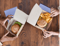 La imagen muestra dos cajas de cartón con comida tipo fast food (comida rápida), colocadas sobre una mesa de madera. En cada caja hay una hamburguesa con pan de sésamo y en una de ellas también hay papas fritas gruesas. Las cajas están siendo sostenidas por las manos de dos personas, una a la izquierda y otra a la derecha.