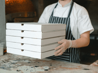 A imagem mostra um profissional de cozinha, com avental listrado e luvas de higiene, segurando uma pilha de caixas de pizza prontas para entrega. O cenário sugere uma operação focada exclusivamente em preparo e despacho de pedidos — sem atendimento ao cliente no local.

Essa é uma representação clássica do funcionamento de uma dark kitchen